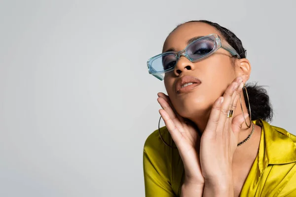 Красива афроамериканська жінка в блакитних сонцезахисних окулярах і стильна сорочка, торкаючись обличчя ізольована на сірому — стокове фото
