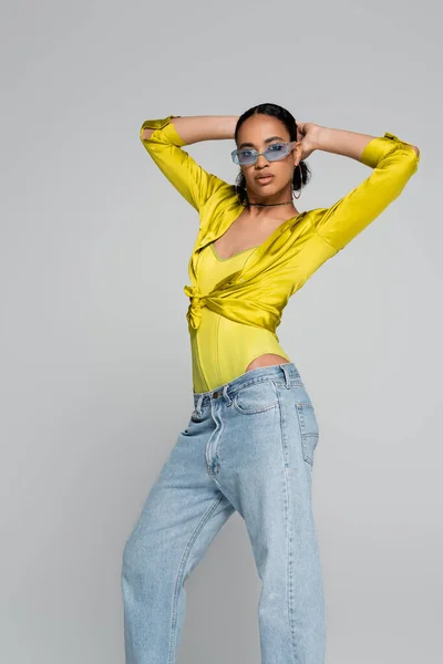 Brünettes afrikanisch-amerikanisches Model mit blauer Sonnenbrille posiert und schaut vereinzelt in die Kamera auf grau — Stockfoto