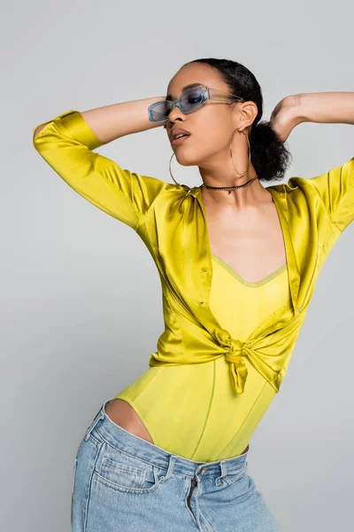 Morena modelo afroamericano en gafas de sol azules posando y mirando hacia otro lado aislado en gris - foto de stock