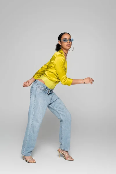 Pleine longueur de femme afro-américaine élégante en lunettes de soleil bleues et vêtements à la mode posant sur le gris — Photo de stock