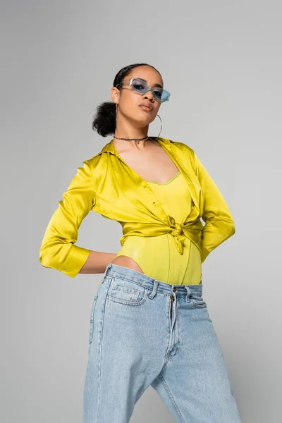 Modèle afro-américain confiant en lunettes de soleil bleues et vêtements tendance posant isolé sur gris — Photo de stock