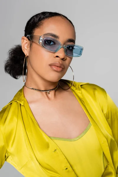 Retrato de la joven modelo afroamericana en gafas de sol azules y ropa de moda posando aislada en gris - foto de stock
