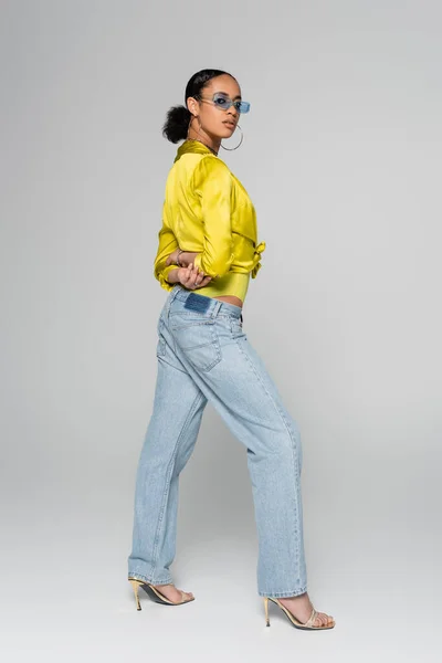 Comprimento total do jovem modelo afro-americano em óculos de sol azuis e roupas da moda posando com as mãos atrás das costas em cinza — Fotografia de Stock