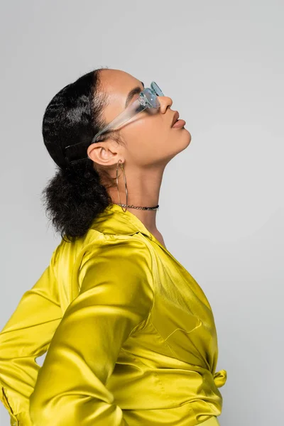 Вид сбоку на африканскую американскую модель в синих солнцезащитных очках и стильную одежду, позирующую изолированно на сером — стоковое фото