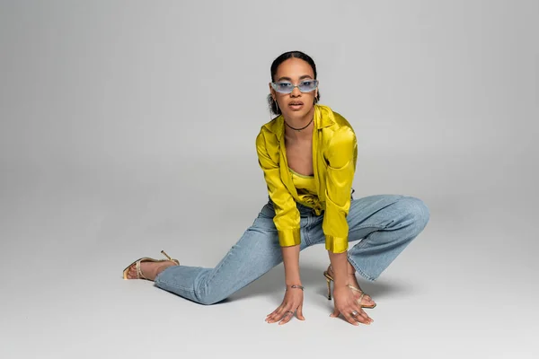 Full length of stylish african american model in blue sunglasses posing while standing on knee on grey background — Stock Photo