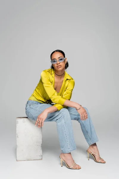 Comprimento total de mulher americana africana elegante em óculos de sol na moda e roupa sentada em cubo de concreto enquanto posando em cinza — Fotografia de Stock