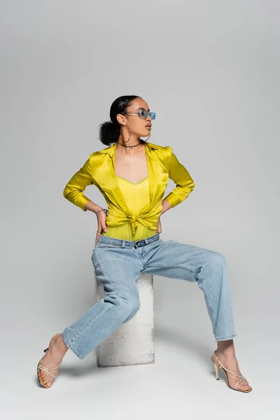 Повна довжина афроамериканської моделі в стильних сонцезахисних окулярах, що сидять на бетонному кубі, позує руками на талії на сірому — стокове фото