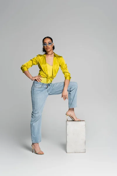 In voller Länge selbstbewusstes afrikanisch-amerikanisches Model tritt auf Betonwürfel, während es mit der Hand auf der Hüfte auf grau posiert — Stockfoto