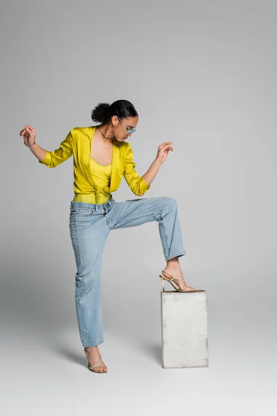 Full length of brunette african american model stepping on concrete cube while posing on grey — Stock Photo