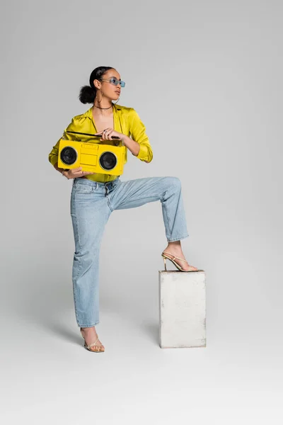 Full length of brunette african american model stepping on concrete cube while holding yellow boombox on grey — Stock Photo