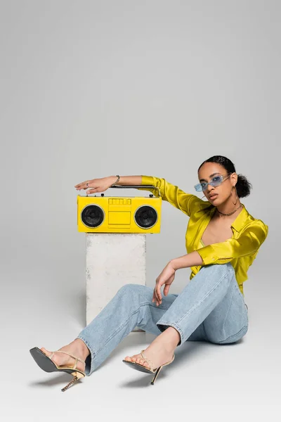 Piena lunghezza di bruna donna africana americana in occhiali da sole seduta vicino al cubo di cemento e retro boombox su grigio — Foto stock