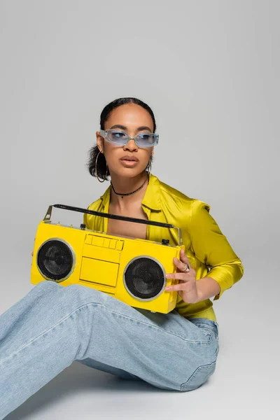 Stylish african american woman in blue sunglasses sitting and holding retro boombox on grey — Stock Photo