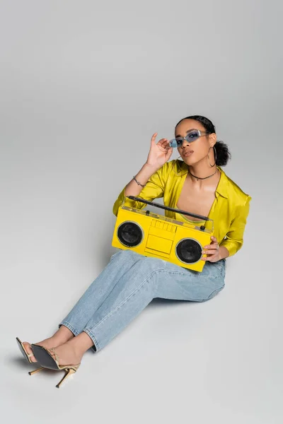 Vue grand angle de femme afro-américaine élégante ajustant les lunettes de soleil bleues et assise avec boombox rétro sur gris — Photo de stock