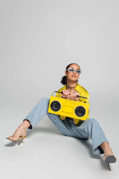 Piena lunghezza di bruna donna africana americana in occhiali da sole blu in posa con boombox giallo su grigio — Foto stock