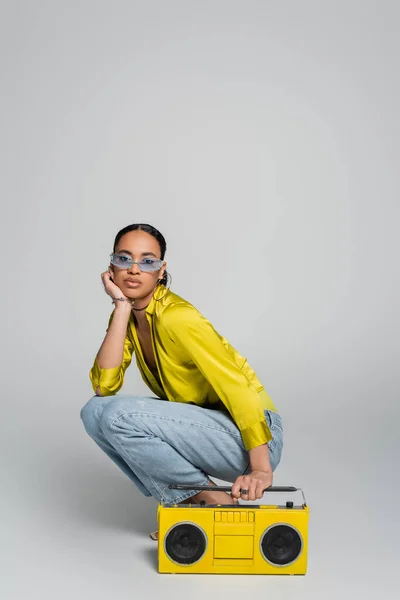 Full length of brunette african american woman in blue sunglasses sitting on haunches near yellow boombox on grey — Stock Photo