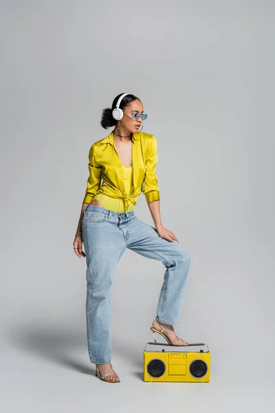 Pleine longueur de femme afro-américaine élégante dans des écouteurs sans fil posant près de boombox jaune sur gris — Photo de stock