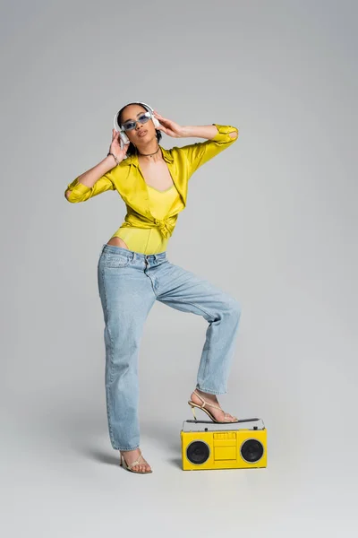 Повна довжина афроамериканської жінки в бездротових навушниках насолоджуючись музикою біля жовтого бумбоксу на сірому — стокове фото