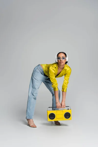 Pleine longueur de femme afro-américaine élégante dans des écouteurs sans fil tenant boombox jaune sur gris — Photo de stock