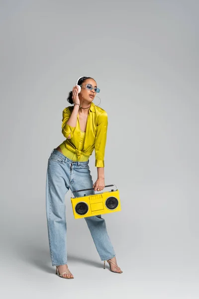 Full length of brunette african american model in wireless headphones holding yellow boombox on grey — Stock Photo