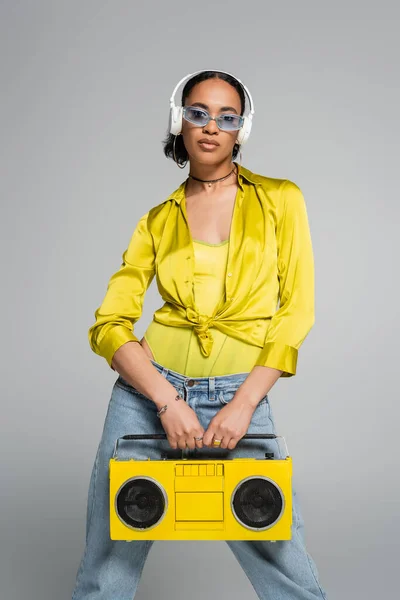 Stylische afrikanisch-amerikanische Frau hört Musik in drahtlosen Kopfhörern und hält Retro-Boombox isoliert auf grau — Stockfoto