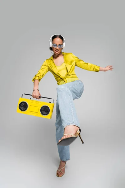 Volle Länge der emotionalen afrikanisch-amerikanisches Modell in drahtlosen Kopfhörern hält gelben Boombox und posiert auf grau — Stockfoto
