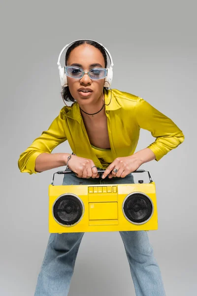 Stylish african american woman listening music in wireless headphones while holding boombox isolated on grey — Stock Photo