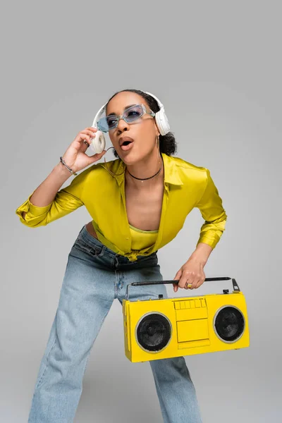 Здивована афроамериканська жінка слухає музику в бездротових навушниках і тримає бумбокс ізольовано на сірому — стокове фото