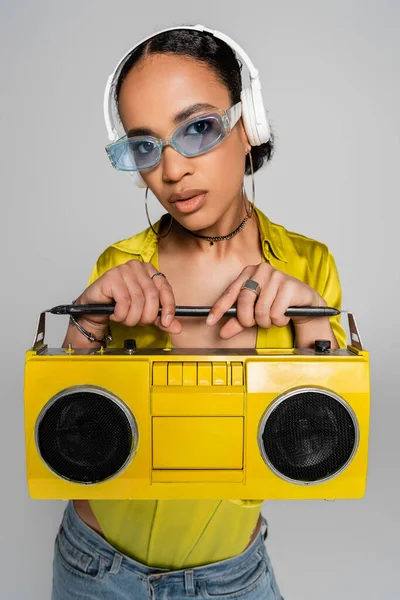 Jolie femme afro-américaine écoutant de la musique dans des écouteurs sans fil et tenant boombox jaune isolé sur gris — Photo de stock