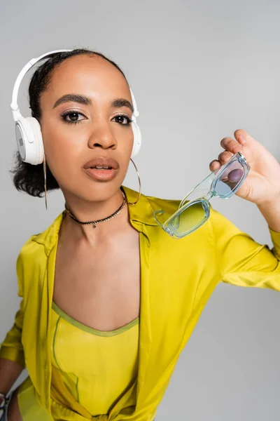 Гарна афроамериканка слухає музику в бездротових навушниках і тримає блакитні сонцезахисні окуляри ізольовані на сірому — стокове фото