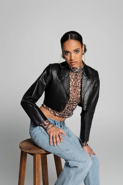 Stylish african american woman in cropped leather jacket and jeans sitting on wooden high chair isolated on grey — Stock Photo