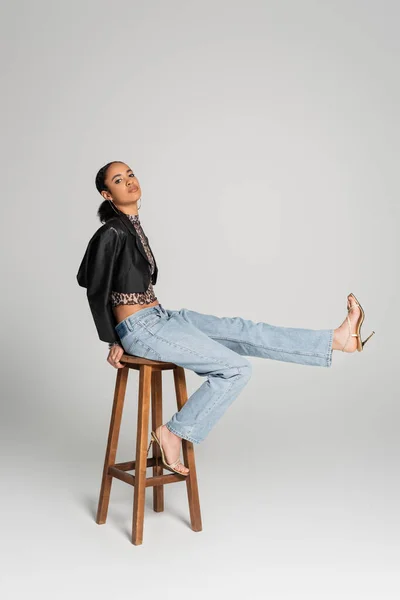 Longitud completa del modelo afroamericano de moda en chaqueta recortada y sandalias de tacón alto sentado en taburete alto en gris - foto de stock