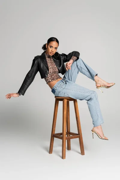 Full length of young african american model in cropped jacket and high heeled sandals posing on high stool on grey — Stock Photo