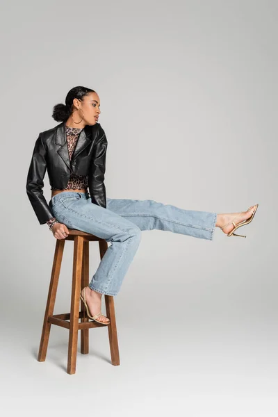 Full length of young african american model in cropped jacket and jeans sitting on high stool on grey — Stock Photo