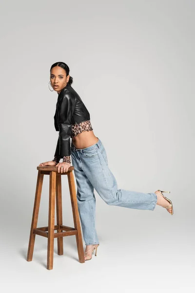 Full length of young african american model in cropped jacket and jeans posing near wooden high stool on grey — Stock Photo
