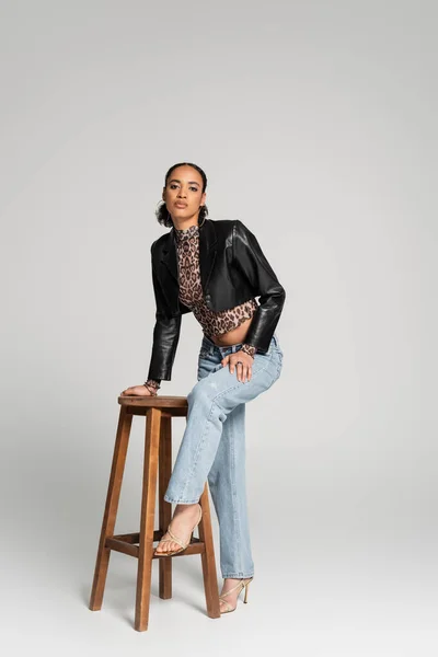 Longitud completa de elegante modelo afroamericano en chaqueta recortada y pantalones vaqueros de pie cerca de la silla alta de madera en gris - foto de stock