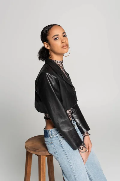 Гарна афроамериканка в обрізаній шкіряній куртці і джинсах, що спираються на дерев'яний стілець ізольована на сірому — стокове фото