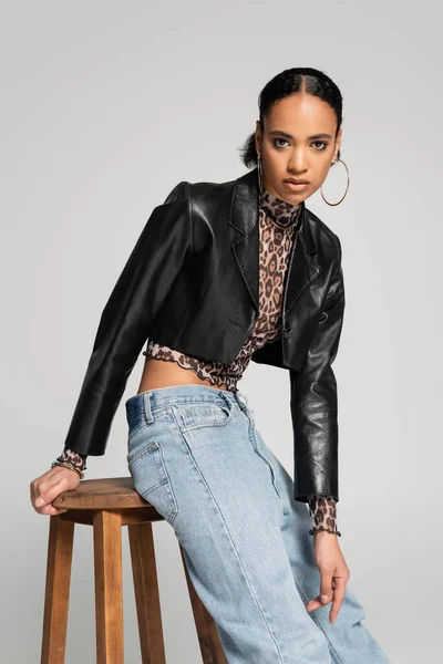 Молодая африканская американская модель в стильной обрезанной куртке и джинсах позирует возле деревянного стула, изолированного на сером — стоковое фото