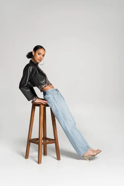 Повна довжина модної афроамериканської жінки в обрізаному піджаку і джинсах, що спираються на дерев'яний високий стілець на сірому — стокове фото
