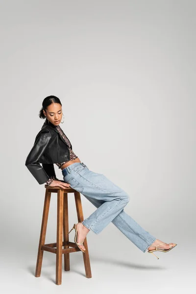 Full length of stylish african american model in cropped jacket and jeans leaning on wooden high chair on grey — Stock Photo