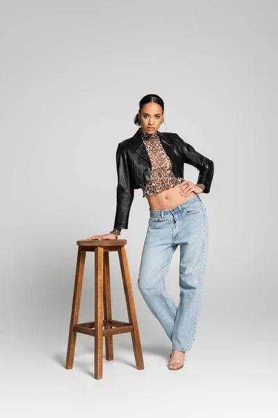 Full length of stylish african american woman in cropped jacket and jeans posing with hand on hip near high chair on grey — Stock Photo