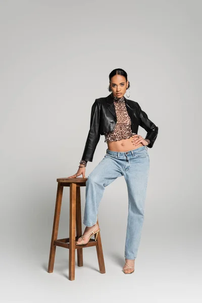 Full length of fashionable african american model in cropped jacket posing with hand on hip near high chair on grey — Stock Photo