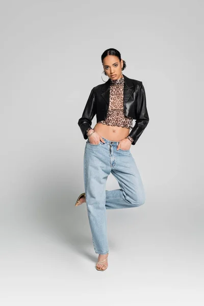 Full length of african american model in cropped jacket posing with hands in pockets while standing on one leg on grey — Stock Photo