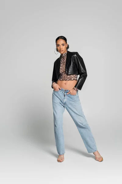 Full length of stylish african american model in cropped jacket and jeans posing with hands in pockets on grey — Stock Photo