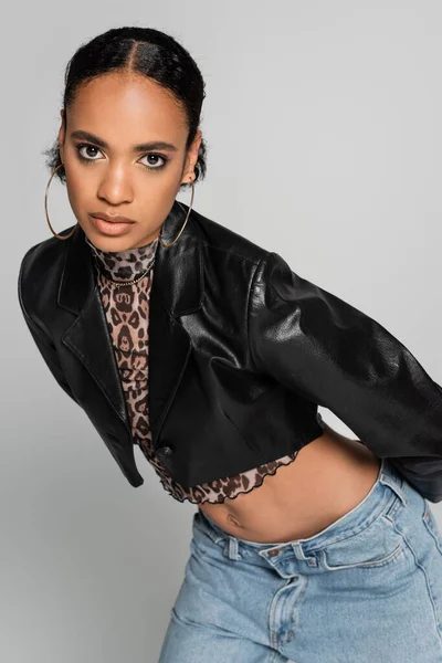 Модна афро-американська модель в обрізаному піджаку і сережках кільця позує ізольовано на сірому — стокове фото
