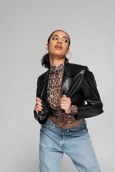 Alla moda donna afroamericana in giacca ritagliata e orecchini a cerchio posa mentre guardando lontano isolato sul grigio — Foto stock