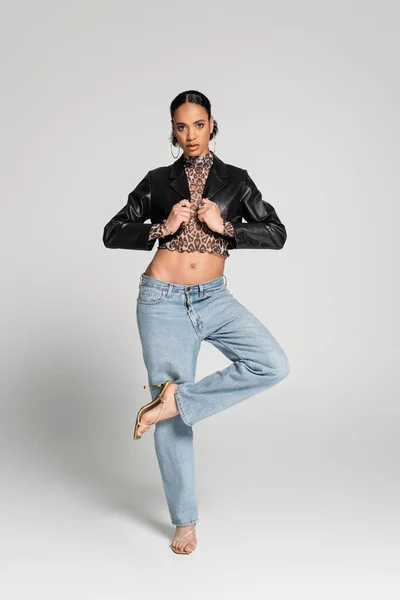 Full length of stylish african american model in cropped jacket and jeans posing on one led while standing on grey — Stock Photo