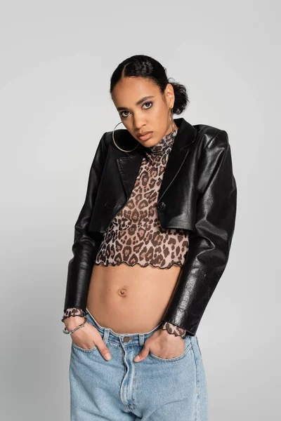 Fashionable african american woman in black cropped jacket and top with animal print posing with hands in pockets isolated on grey — Stock Photo