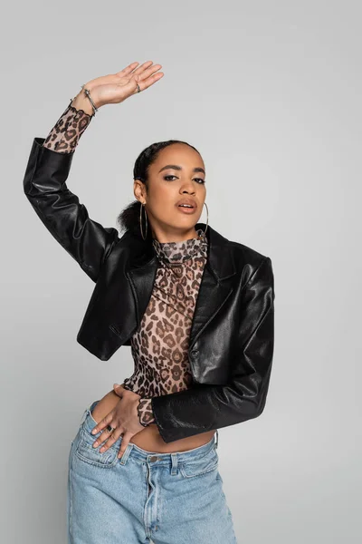 Fashionable african american woman in black cropped jacket and top with animal print posing with raised hand isolated on grey — Stock Photo