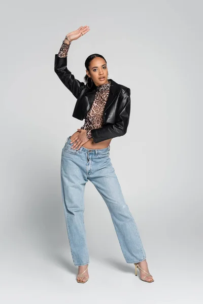 Full length of fashionable african american woman in black cropped jacket posing with raised hand on grey — Stock Photo