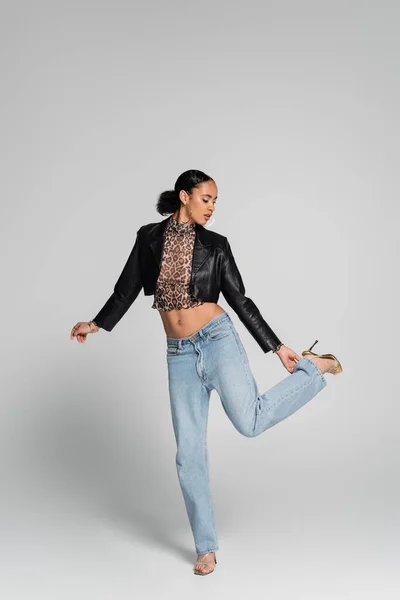 Comprimento total da mulher americana africana elegante em saltos altos de pé uma perna e ajustando jeans em cinza — Fotografia de Stock